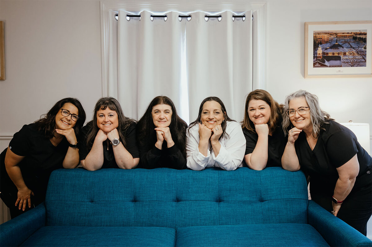 Photo of the six women who make up the Clearwater Family Dental team. They are posed behind a blue sofa.
